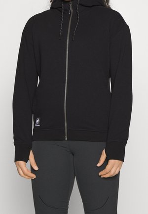 Volwassene draagt een zwarte zip-hoodie met trekkoorden en donkergrijze broek, staand tegen een effen lichte achtergrond.