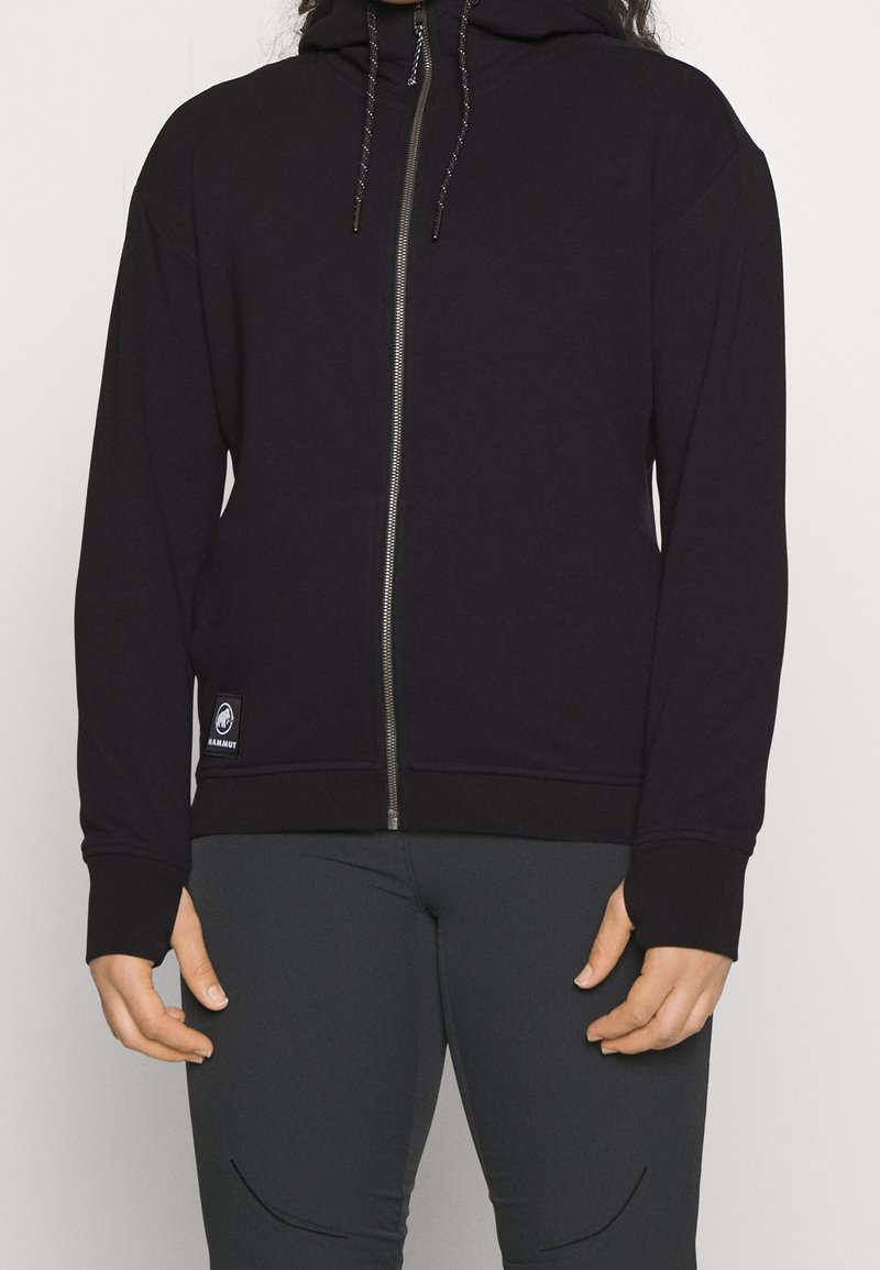 Adulte portant un sweat à capuche noir zippé avec cordons et un pantalon gris foncé, debout devant un fond clair uni.
