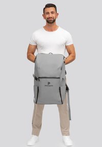 Sac à dos gris au design épuré, avec de multiples compartiments, des bretelles réglables et un logo à l'avant. Texture lisse et forme fonctionnelle.
