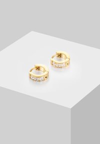 Zelta auskari ar trim skaidrām dārgakmeņiem katrā, novietoti uz balta pjedestāla gaiši pelēkā fona.