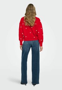 Červený pletený svetr s květinovou výšivkou, kombinovaný s modrými širokými džínami s vysokým pasem. Měkká textura, volný střih, žebrované manžety a lem.