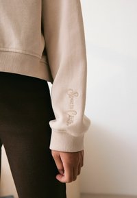 Beżowy sweter z prążkowanymi mankietami, z haftowanym logo "Sports Club" na rękawie. Noszony z ciemnymi prążkowanymi legginsami.