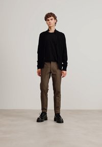 Svart zip-up jakke over en svart t-skjorte, sammen med teksturerte brune bukser og kraftige svarte sko. Minimalistisk stil mot en lys bakgrunn.