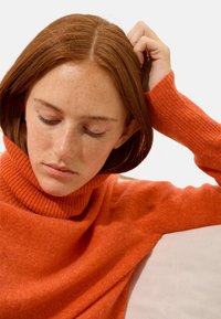 Oranges Rollkragenpullover mit geripptem Muster, der elegant fällt. Der Fokus liegt auf dem weichen Stoff und dem lockeren Schnitt, mit einem subtilen Glanz.