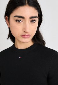 Svart ribbestrikket genser med rund hals, med en liten logo i rødt, hvitt og blått på brystet, glatt tekstur.