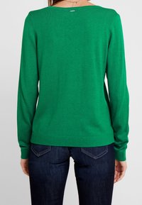 Grüner Langarmpullover mit rundem Ausschnitt, strukturierter Rippoptik und einem kleinen Etikett auf der Rückseite. Kombiniert mit dunkler Jeans.