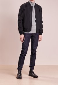 Mann trägt eine schwarze Bomberjacke, einen grauen Reißverschluss-Pullover, dunkelblaue Jeans und schwarze Lederstiefel, steht auf einem beigen Boden vor einer hellen Wand.