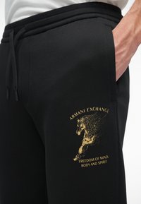 Crne trenirke s podesivim pojasom, koje s lijeve strane bedra imaju zlatni grafik s konjem i tekst "Armani Exchange". Glatka tekstura.
