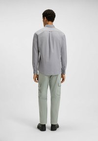 Camisa gris de manga larga con cuello, detalle de pliegue trasero, combinada con pantalones cargo verde claro y zapatos negros, vista desde la parte de atrás.