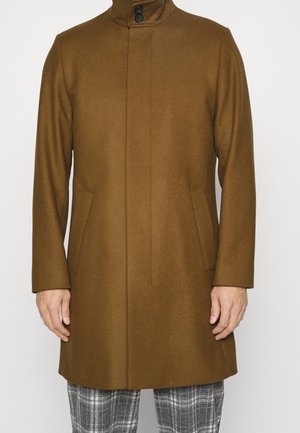 Abrigo de lana marrón con cuello alto y cierre de botones, llevado sobre pantalones de cuadros grises, mostrado desde la mitad del muslo hasta el cuello.