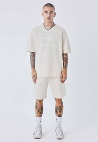 Crème t-shirt met korte mouwen en een wit logo, gecombineerd met bijpassende shorts. Sneakers en sokken met zichtbare ontwerpen. Getatoeëerde armen.