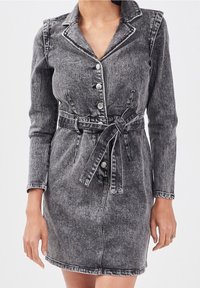 Robe en denim noir délavé avec manches longues, coupe ajustée, col, boutonnage sur le devant et ceinture en tissu. Présente des coutures contrastées et une texture subtile.