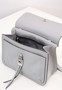 Šedá kožená kabelka s texturovaným povrchem, obdélníkovým tvarem, klopovým uzavřením a malým stříbrným kovovým detailem. Uvnitř je zipová kapsa.