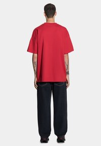 Czerwona bawełniana koszulka z krótkim rękawem o luźnym kroju, pokazana z tyłu. Posiada okrągły dekolt i prosty fason. W połączeniu z ciemnoniebieskimi dżinsami.