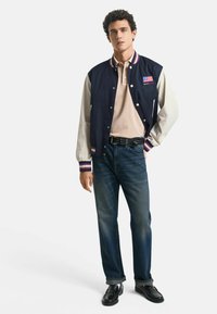 Marineblå varsityjacka med krämfärgade ärmar, röd och blå randig krage, beige polotröja, blå jeans, svarta skor. Har logotyp på bröstet.