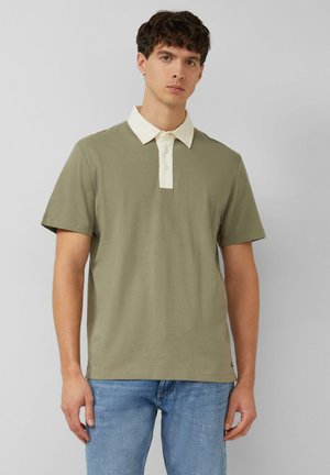 Giovane uomo che indossa una polo verde oliva a maniche corte con colletto bianco e jeans blu, in piedi contro uno sfondo chiaro e uniforme.