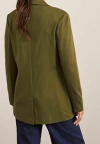 Olijfgroen op maat gemaakte blazer met een enkele split en lange mouwen, vervaardigd uit textuurstof. Gecombineerd met donkerblauwe wijde broek.
