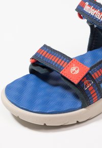 Blå sandal med gummisula, textilremmar med röda och blå mönster, justerbara stängningar och en logotyp på sidan.