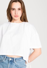 Camiseta branca corta com decote redondo e mangas curtas, feita de tecido leve, com laços laterais e um corte descontraído.