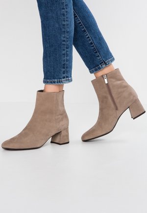 Beżowe zamszowe botki za kostkę na szerokim obcasie, z bocznym zamkiem błyskawicznym i zaokrąglonym noskiem. Noszone z podwiniętymi niebieskimi jeansami.