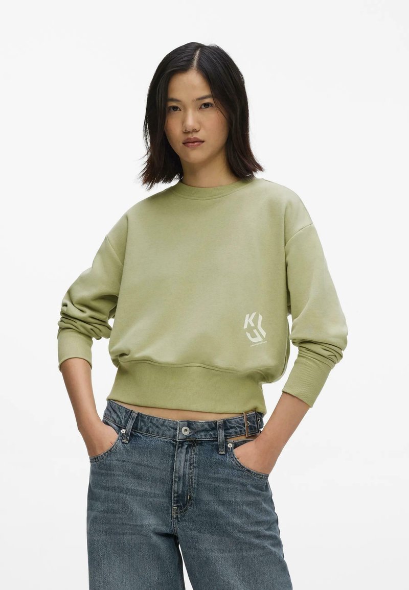 Femme aux cheveux foncés mi-longs portant un sweat-shirt court vert clair et un jean bleu, les mains dans les poches.
