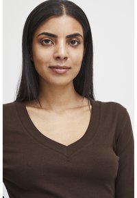 Braunes, geripptes Langarmoberteil mit V-Ausschnitt. Der Stoff wirkt weich und hat eine taillierte Silhouette, die den Ausschnitt und die Schultern betont.