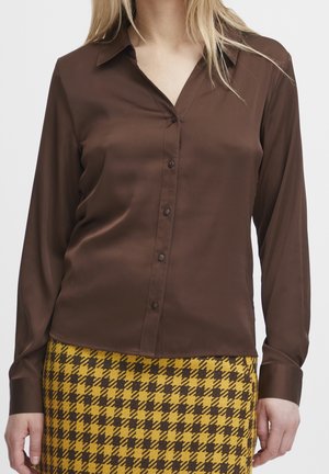 Blouse marron à manches longues avec un col et une fermeture à boutons, fabriquée en tissu lisse. Associée à une jupe à carreaux jaune et marron.