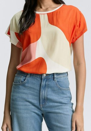 Kobieta ubrana w krótki rękaw z abstrakcyjnym wzorem w kolorach pomarańczowym, kremowym i różowym, w zestawieniu z wysokim stanem niebieskich dżinsów.