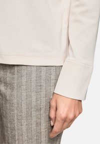Light beige long-sleeve top with a rounded hem. Features subtle stitching and a "Marc Cain" signature at the bottom. Paired with light gray pinstripe pants.