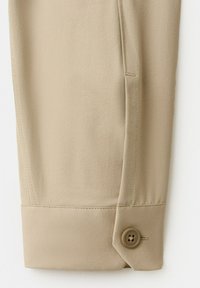 Pied de pantalon beige avec un détail de patte boutonnée cousue près de l'ourlet sur un fond blanc uni.