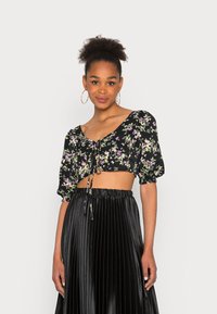 Top cropped preto com padrão floral, mangas curtas em balão e laço à frente. Combinado com uma saia preta plissada, criando um visual texturizado.