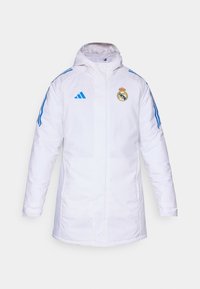 Hvid isoleret jakke med hætte, der har blå striber og Real Madrid-logoet på brystet. Fremstillet af glat, vejrbestandigt materiale.