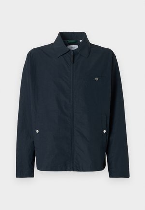 Giacca leggera blu navy con colletto appuntito, cerniera frontale completa e una tasca sul petto. Presenta dettagli con bottoni a pressione sulle tasche.