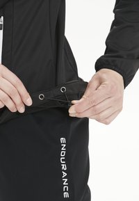 Sort atletisk jakke med snøre i taljen og lynlås. Stoffet ser glat ud. Hånd justerer snøret. Logo synligt på bukserne.