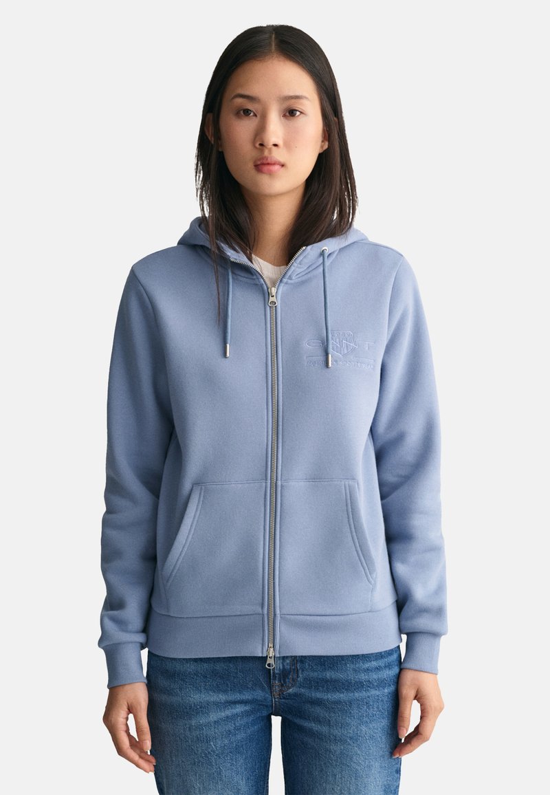 Lichtblauwe zip-up hoodie met een capuchon, ribbelkraag, een voorzak en geborduurd logo op de borst. Zachte fleecemateriaal en een relaxte pasvorm.