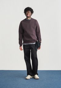 Σκούρο μοβ φούτερ με ριμπ μανσέτες, σε συνδυασμό με μαύρο παντελόνι. Λευκό πουκάμισο φορεμένο από κάτω· μαύρο καπέλο κρεμασμένο στην τσέπη του παντελονιού. Μπεζ αθλητικά παπούτσια.