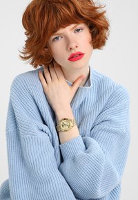 Suéter de punto azul claro con corte holgado y textura acanalada, combinado con un reloj dorado con esfera redonda y tres subesferas.