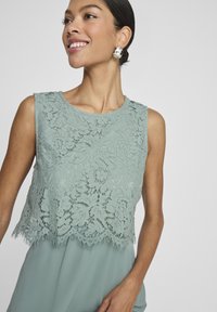 Blonde topp i myk grønn med blomstermønster, med bølgekant og frynsete kanter. Ermeløs design med rund hals.