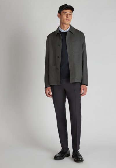 Donkergrijs, op maat gemaakte jas met een kraag, knoopsluiting en twee vo zakken, gecombineerd met donkere broeken en zwarte schoenen.