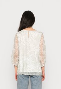 Blusa transparente com um leve padrão floral em suaves tons de branco e bege, apresentando mangas três quartos e um detalhe em forma de buraco na parte de trás.