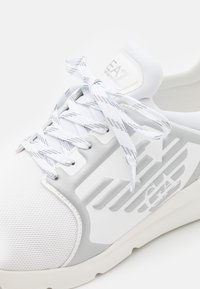 Białe i jasnoszare sneakersy z wzorzystymi sznurówkami, siateczkowym czubkiem oraz logo EA Emporio Armani z boku i na języku.