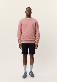 Ανοιχτό ροζ φούτερ με στρογγυλή λαιμόκοψη, φορεμένο πάνω από λευκό μπλουζάκι, σετ με ναυτικομπλε σορτς και μπλε αθλητικά παπούτσια, συμπληρωμένο με γκρι κάλτσες.