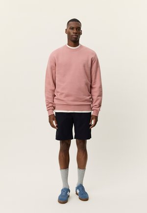 Vaaleanpunainen collegepaita pyöreällä kaula-aukolla, valkoisen t-paidan päällä, yhdistetty laivastonsinisiin shortseihin ja sinisiin lenkkareihin, joita täydentävät harmaat sukat.