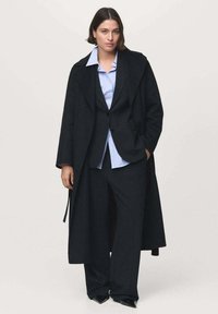 Manteau long en mélange de laine noire superposé sur un costume rayé noir et une chemise légère bleu clair. Comprend un col pointu et des chaussures noires élégantes.