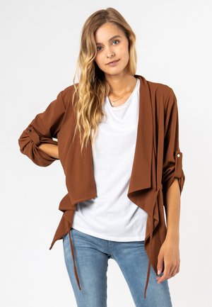 Bruine, lichte open voorloper cardigan met opgerolde mouwen en strikdetails. Gekocht over een witte T-shirt en gecombineerd met lichtblauwe spijkerbroek.