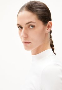 Witte geribde coltrui met een aansluitend ontwerp. De persoon heeft bruin haar in een vlecht en draagt kleine stud oorbellen.