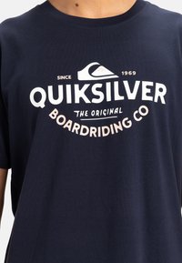 Camiseta de manga corta de color azul marino hecha de algodón, con un logotipo gráfico en blanco y rosa que presenta un diseño de ola y detalles en texto.