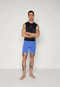 Joven con tatuajes que lleva una camiseta sin mangas negra de Nike y pantalones cortos brillantes azules de Nike, de pie descalzo con los brazos cruzados frente a un fondo liso.