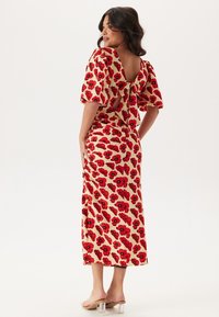 Robe midi fleurie en tissu beige avec imprimé coquelicot rouge, présentant des manches ballons, un dos nu avec des liens, et une silhouette ajustée.