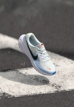 Nike løbesko i lyseblåt netmateriale med sort swoosh, orange accenter og en tekstureret sål. Hævet over en malet hvid linje.